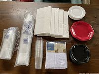 Image showing white disposable plastic cups, tall white paper napkins, clear plastic drinking cups, white paper plates, red plastic octagonal plates, black plastic round plates arranged on a wooden surface.