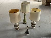 Four pottery based lamps arranged on a floor, two beige and brown matching lamps with shades, one vintage green lamp with scalloped lampshade, one small dark brown lamp base without shade.