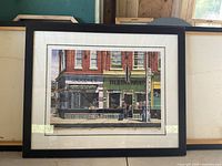 Framed limited edition print showing a street scene of Queen Street West at Trinity, Toronto, featuring a grocery and restaurant storefront with architectural details in a black frame.