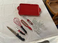 Red rectangular Tara casserole tray with wavy design, three clear faceted glass tea light holders, and various kitchen utensils including knives, whisks, and grater on white cloth.