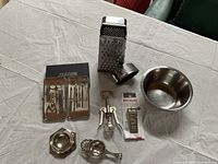 Photo showing silver manicure set in original blister packs alongside metal box grater shredder, stainless steel bowl, winged corkscrew, tea bag holder, and small round metal ashtray or dish all placed on white fabric surface.