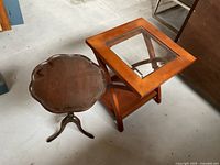 Photo showing square wooden side table with glass top and round wooden pedestal table side by side
