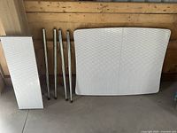 White Formica tabletop with silver metal legs arranged beside it on a concrete floor in front of a wooden wall.