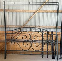 Black wrought iron bed frame headboard and footboard with scroll and geometric designs, standing against a wooden and metal warehouse wall