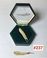 Gold encrusted goose feather brooch inside hexagonal green box with feather illustration on lid, and second brooch outside box on white background