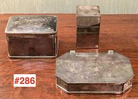 Three silver plated dresser boxes shown together on a wooden surface. Boxes are tarnished with visible patina, showing typical age wear.