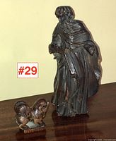 Side view of the 19th century wooden religious figure carving showing detailed robe folds and stance against a neutral background on a wooden surface with a smaller carved animal figure.