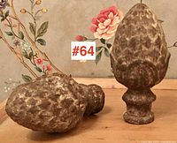 Two antique cast iron pinecone shaped gate post toppers on wooden surface, one standing upright and one lying on its side showing hollow base.