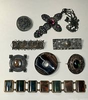 All eight items arranged on white background: round brooch, cross pendant, two bar brooches, square brooch, circular agate brooch, oval brooch, agate panel bracelet