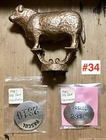 Front view of gold-tone metal Dodge cow/bull ornament with textured details and lower ornate base showing threaded screw attachment, accompanied by two sealed 1940s Day Deck metal ID tags labeled PASCOL with numbers 2310 and 2319.