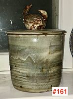 Front view of cylindrical pottery canister with layered green and brown glaze, featuring a bird figurine lid.
