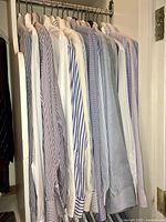 Side angle of 18 men's dress shirts hanging on rack, displaying variations of stripes, checks, and solid colors in blue, white and gray shades.
