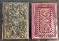Top view of two antique books with decorative leather bindings showing intricate floral patterns in gold and green on brown and red covers.