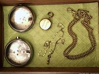 Drawer display showing two open pocket watch cases at left, small round fob at top center, geometric link chain in middle, curb link chain at right