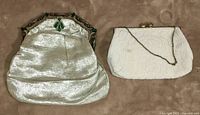 Two vintage purses side by side on beige fabric; left is silver mesh with green accents, right is white beaded with gold-tone chain.