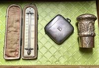 Three antique items: cased thermometer on left, sterling vesta case in middle, ornate walking stick handle on right, displayed on green surface.