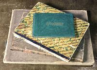 Four vintage books stacked on a surface; the top book is an autograph album with dark green embossed cover and gold lettering 'AUTOGRAPHS'. The other books have marbled and textured covers with wear.