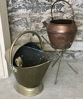 Two antique metal vessels: a brass bucket and a copper watering can on a tripod stand, both showing patina.