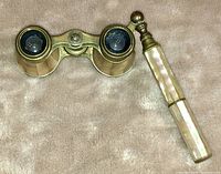 Close-up of vintage opera glasses with mother of pearl and brass detailing on a beige fabric background.