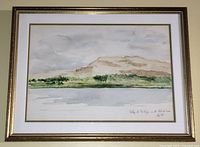 Full frontal image of framed watercolour landscape with gold frame and white matting. Scene shows distant mountains, trees and water under cloudy sky.