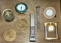 Assorted desk drawer items arranged on a wooden surface showing three small clocks, two large brass medallions, a brass cup, metal stapler, and a large metal nail.