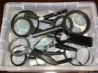 Photo showing a container filled with a variety of magnifying glasses with black handles in different sizes and styles.