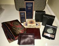 Wide view of the lot showing all items: L'Occitane shaving products in original boxes (brush, razor, soap), two wallets, Pierre Cardin silk scarf, and a black leather pouch with magnifier.