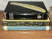 Four books stacked on a wooden surface: large black folio with a curved ivory artifact on cover, Mythology book with gold letters, A Catalogue of Renaissance Medals with blue cover dust jacket, black cover edges visible.