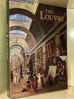 Front cover of The Louvre hardcover coffee table art book showing artwork of the museum's interior gallery with people and statues.