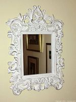 Full view of the ornate white carved mirror hanging on a wall, showing the entire frame and mirror surface reflecting part of a room with framed artwork.