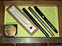 Three fountain pens on quilted mat with wood-framed thermometer and brass medallion