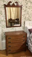 Full view of mahogany chest of drawers with attached mirror above, set against floral wallpaper and beside a bed