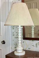 Full view of the white ceramic table lamp with a pleated cream lampshade on a wooden surface, reflecting in a mirror behind.