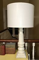 Full view of the vintage white distressed table lamp on a yellow table near a railing, showing the lamp base and white drum shade.
