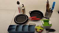 Overview of kitchen items on table showing pans, skillet, water bottles, utensil holder with kitchen tools, food containers, and accessories.