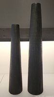 Two tall black vases shown side by side against a neutral backdrop with soft light highlighting their texture and form.