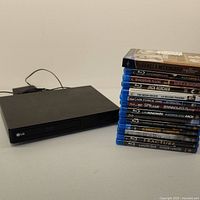 Photo showing black LG Blu-ray player unit with power cable and stack of 15 Blu-ray and DVD movie cases on a flat surface.