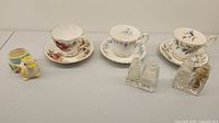 Photo shows three fine china tea cups and saucers with floral designs, two pairs of glass salt and pepper shakers, and a small colorful ceramic bird-shaped salt and pepper shaker set.
