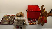 Photo of assorted Christmas decorations including Villeroy & Boch manger scene, ornaments stored in multi-compartment fabric storage boxes, a red fabric box, plaid gift box covers, a wooden reindeer, and tealight candles.