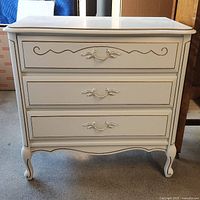 Front view of the white wooden dresser showing three drawers and gold trim decorative handles and outlines.