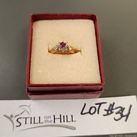 Top view of the gold ring in a cream-colored ring box showing the red gemstone surrounded by cubic zirconia.