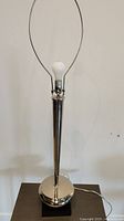 Full view of the lamp without shade, showing tall chrome tapered body, circular base on black square stand, with light bulb and harp fitting on top.