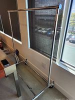 Full view of a single bar metal clothing rack with chrome finish positioned near a window, showing the caster wheels at the base and the collapsible frame structure.