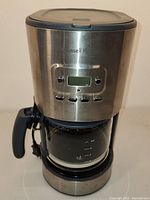 Front view of Russell Hobbs coffee maker showing stainless steel body, digital display, buttons, and glass carafe with handle.