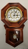 Front view of the full vintage wooden wall clock mounted on wall, octagonal shape with lower pendulum compartment visible.