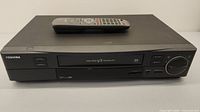 Front view of Toshiba VHS player with remote resting on top, showing front controls and cassette slot.