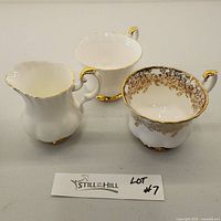 Three pieces of Royal Albert Val D'or china - a tea cup with gold floral pattern and rim, white creamer with gold trim, and sugar bowl with gold rim and floral pattern inside.
