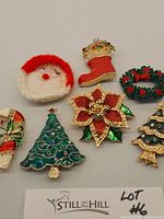 Seven vintage Christmas-themed costume jewelry pins arranged on a plain background: Santa face with yarn hair and nose, red stocking with crown and rhinestones, green wreath with red berries, poinsettia flower with red glitter petals, two Christmas tree pins with rhinestones, and a candy cane pin with enamel and rhinestones.