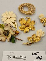 Five vintage costume jewelry brooches arranged on a surface: a cream dogwood flower, cream enamel daisy, textured gold-tone coiled spiral, gold-tone rose flower, and a pair of cream rose buds.