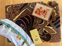 1970's orange and brown shag hooked rug, partially folded; framed Holly Hobbie embroidery art, fabric with floral and figure motifs in warm tones; section of white curtain with colorful embroidered animal patterns; vintage baby food warmer original box; vintage first aid kit box